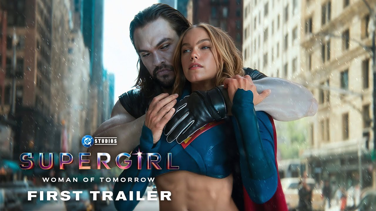 A woman in a blue superhero suit stands beside a man in dark armor; “Supergirl: Woman of Tomorrow” and “First Trailer” text.
