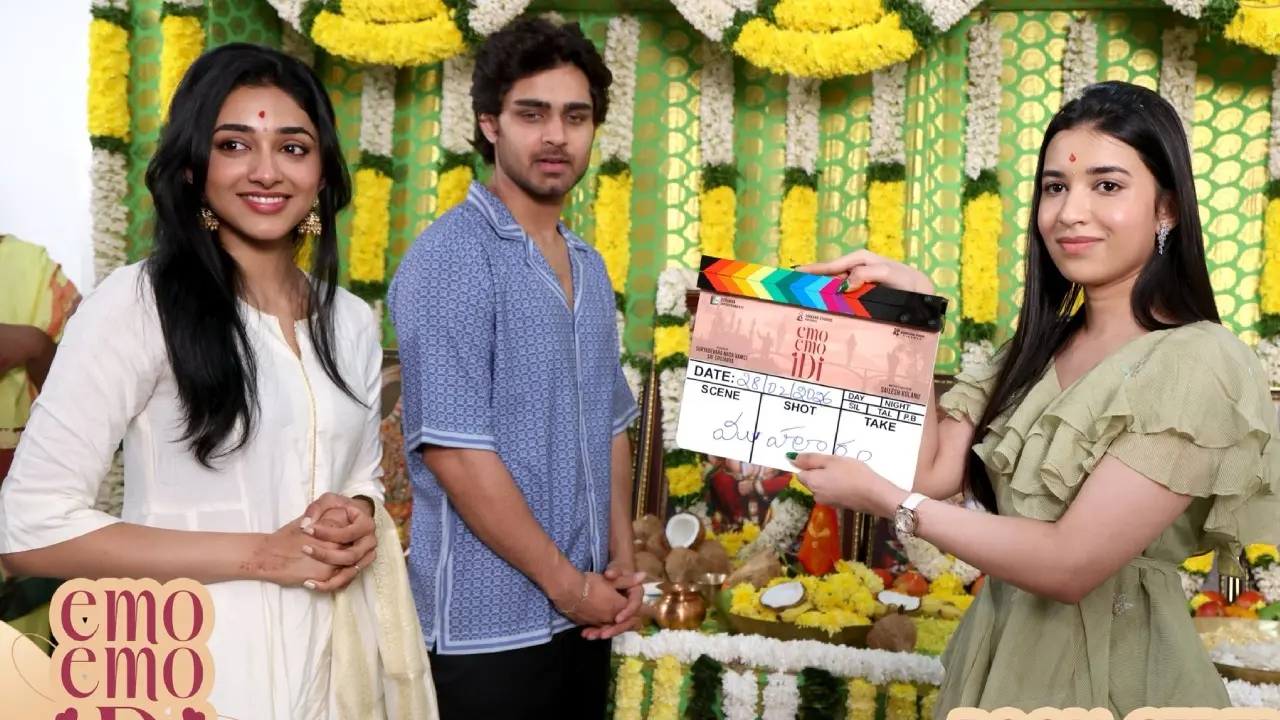 Three people pose at a flower-decorated film set as a woman holds an Emo Emo Idi clapperboard.