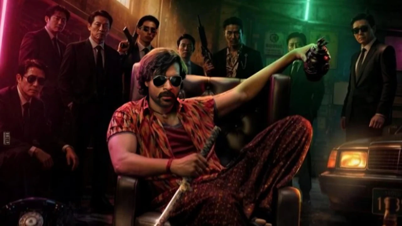 A man in sunglasses and a red patterned outfit sits amid several suited men in a dim, color-lit room.