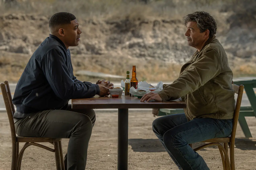 Two men sit across from each other at an outdoor table in Lanterns.