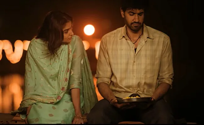 A woman in a green sari looks toward a man holding a plate under warm lights.
